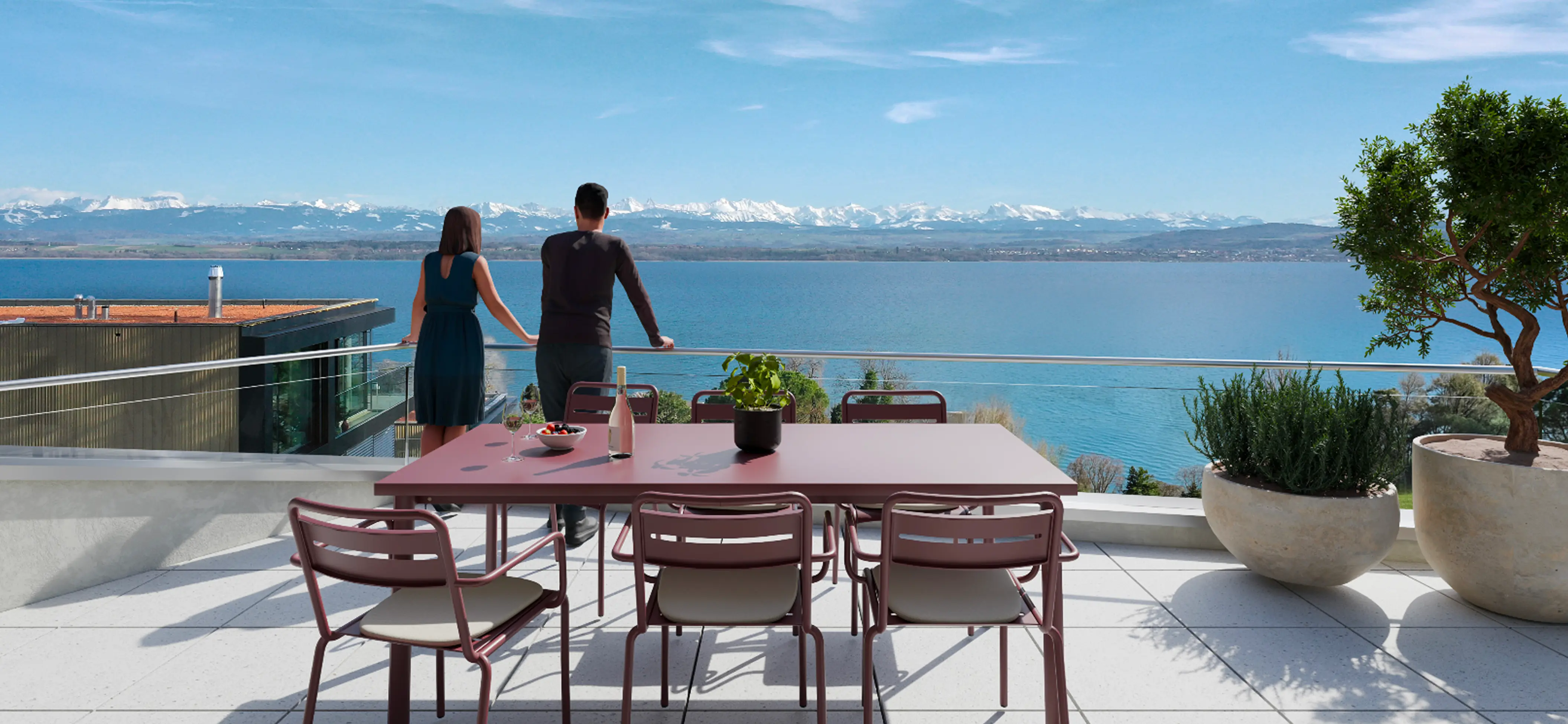Couple debout sur une terrasse moderne avec table et chaises, admirant un lac bleu et des montagnes enneigées à l'horizon.