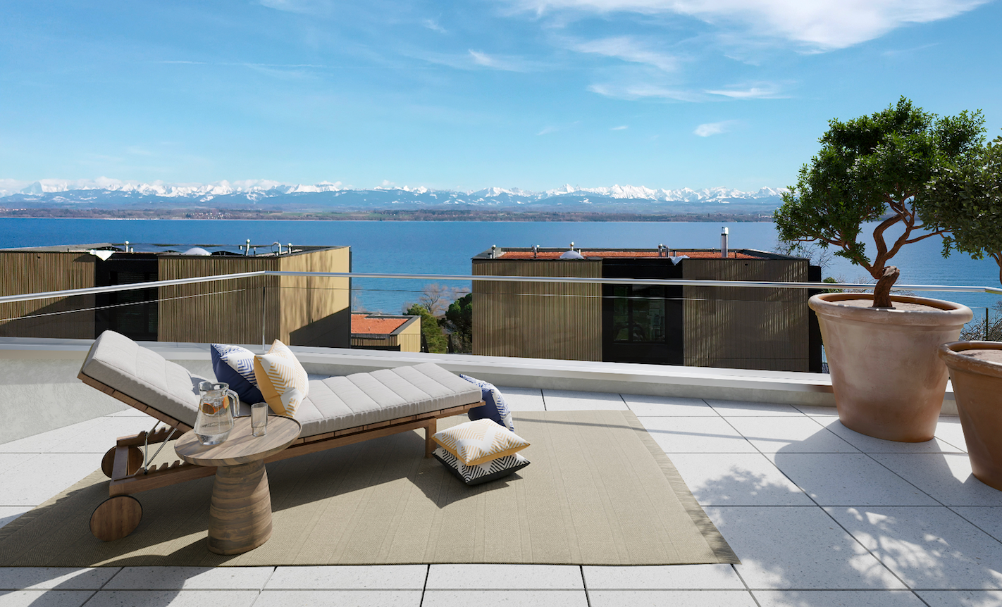 Terrasse avec une chaise longue gris clair et une petite table en bois, vue sur un lac bleu et des montagnes enneigées en arrière-plan.