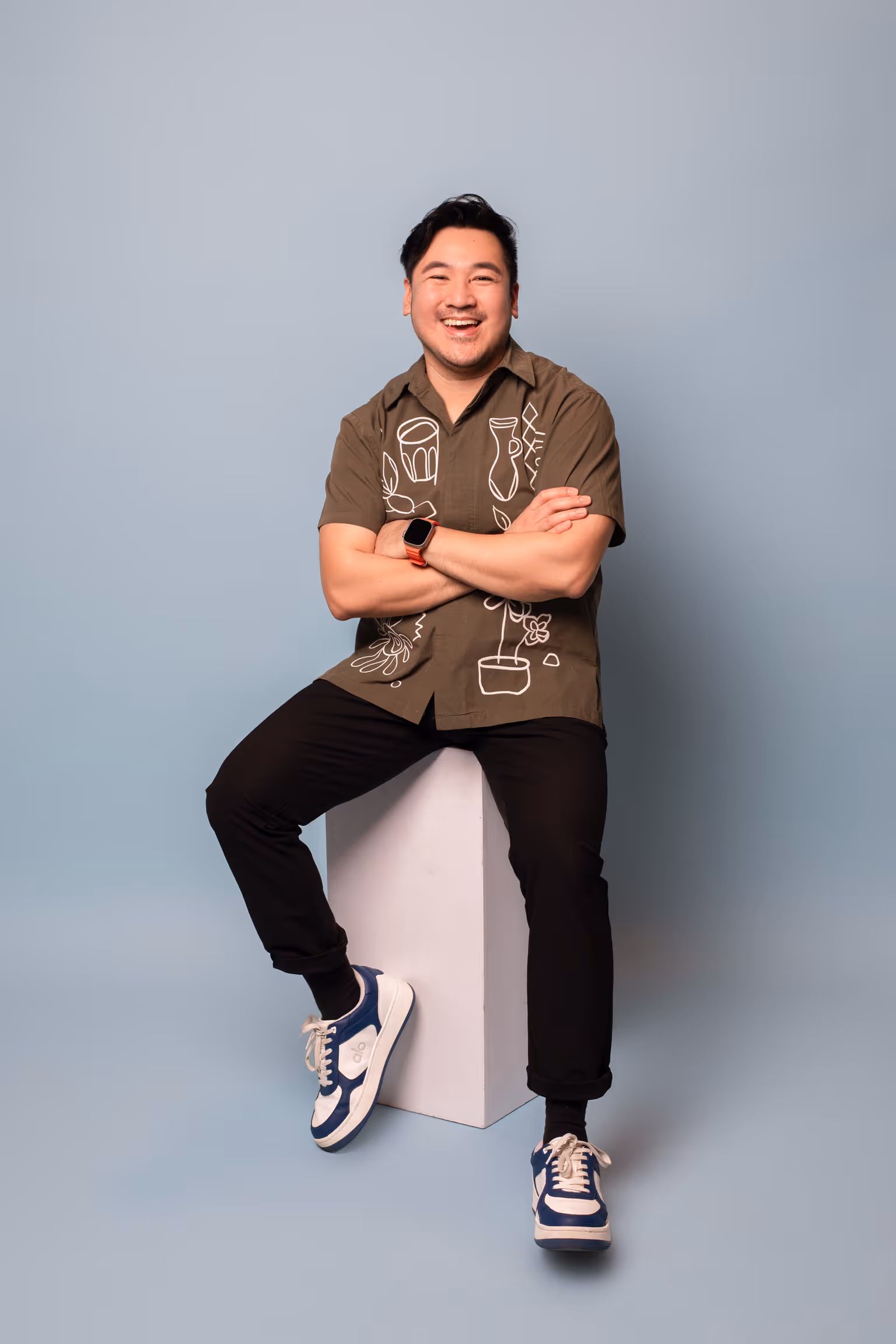 Smiling man sitting on a white block against a light blue background wearing a brown shirt with white line art designs, black pants, and blue and white sneakers.