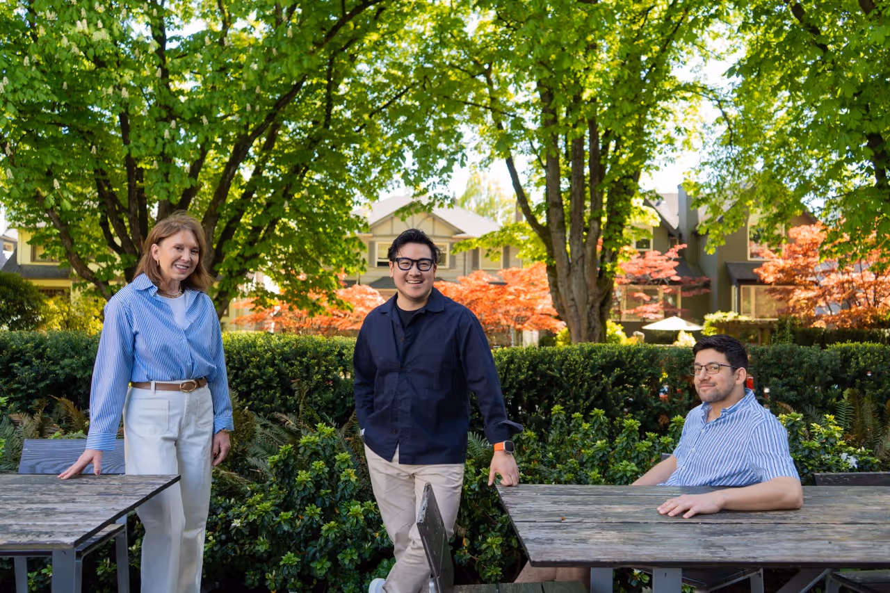 Three people smiling outdoors near wooden tables with green trees and houses in background.