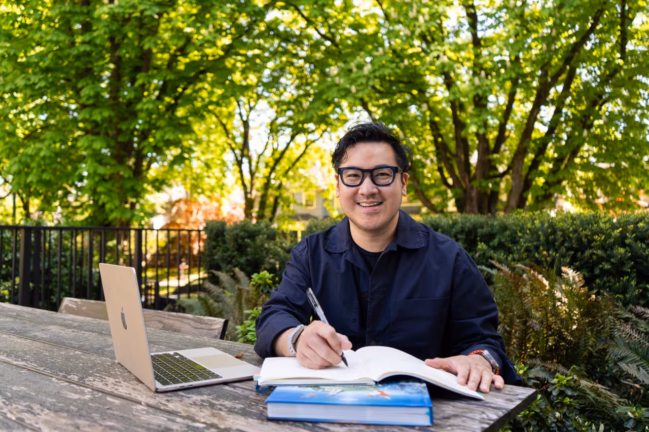 Man with glasses smiling while writing in a book at an outdoor wooden table with a laptop nearby.