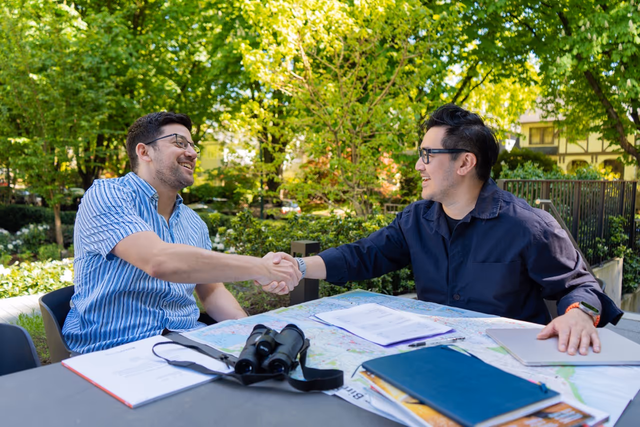 Two men outdoors shaking hands across a table with papers, binoculars, and notebooks.