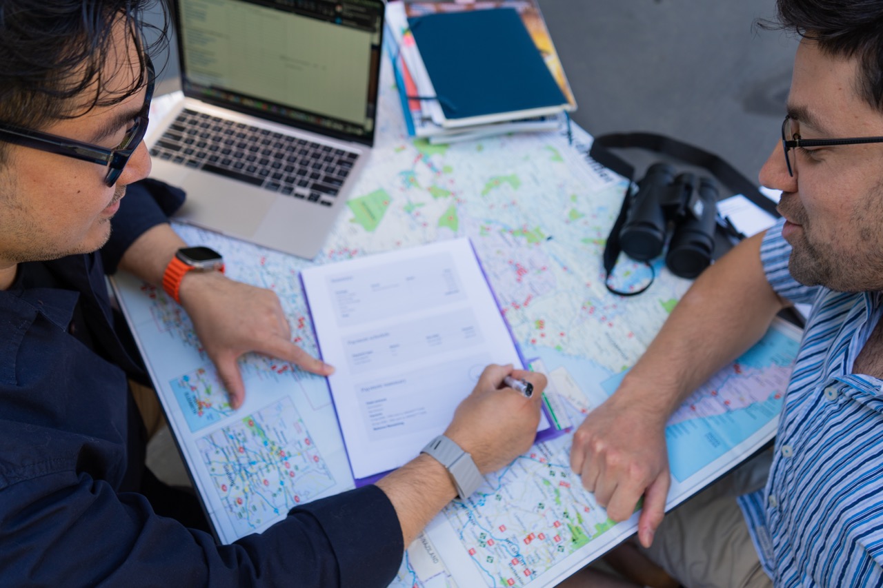 Two men planning a trip with maps, notebooks, a laptop, and binoculars on a table.