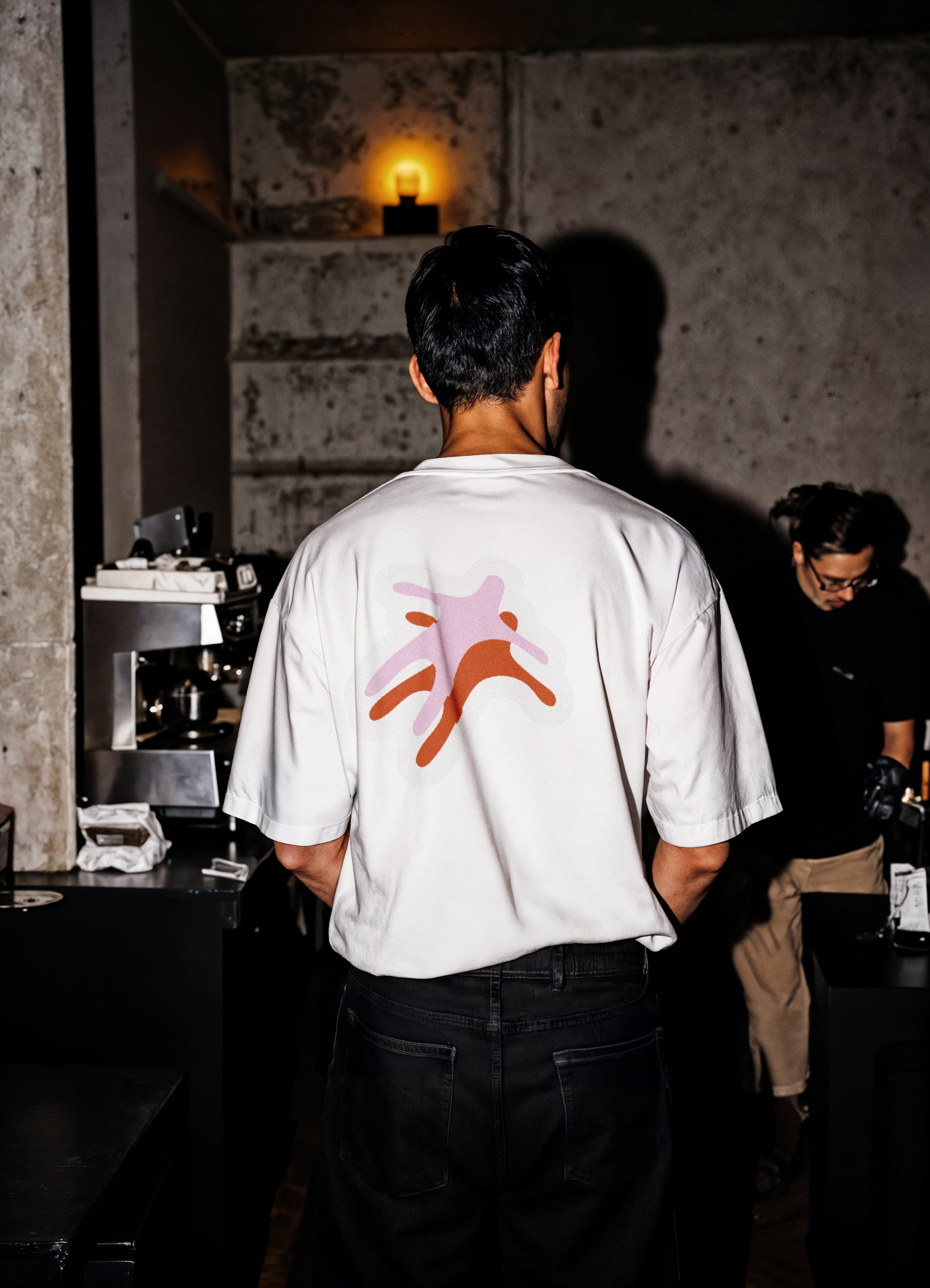 mockup found on magnific -Man wearing white t-shirt with abstract pink and red design on back, standing indoors in dim light.
