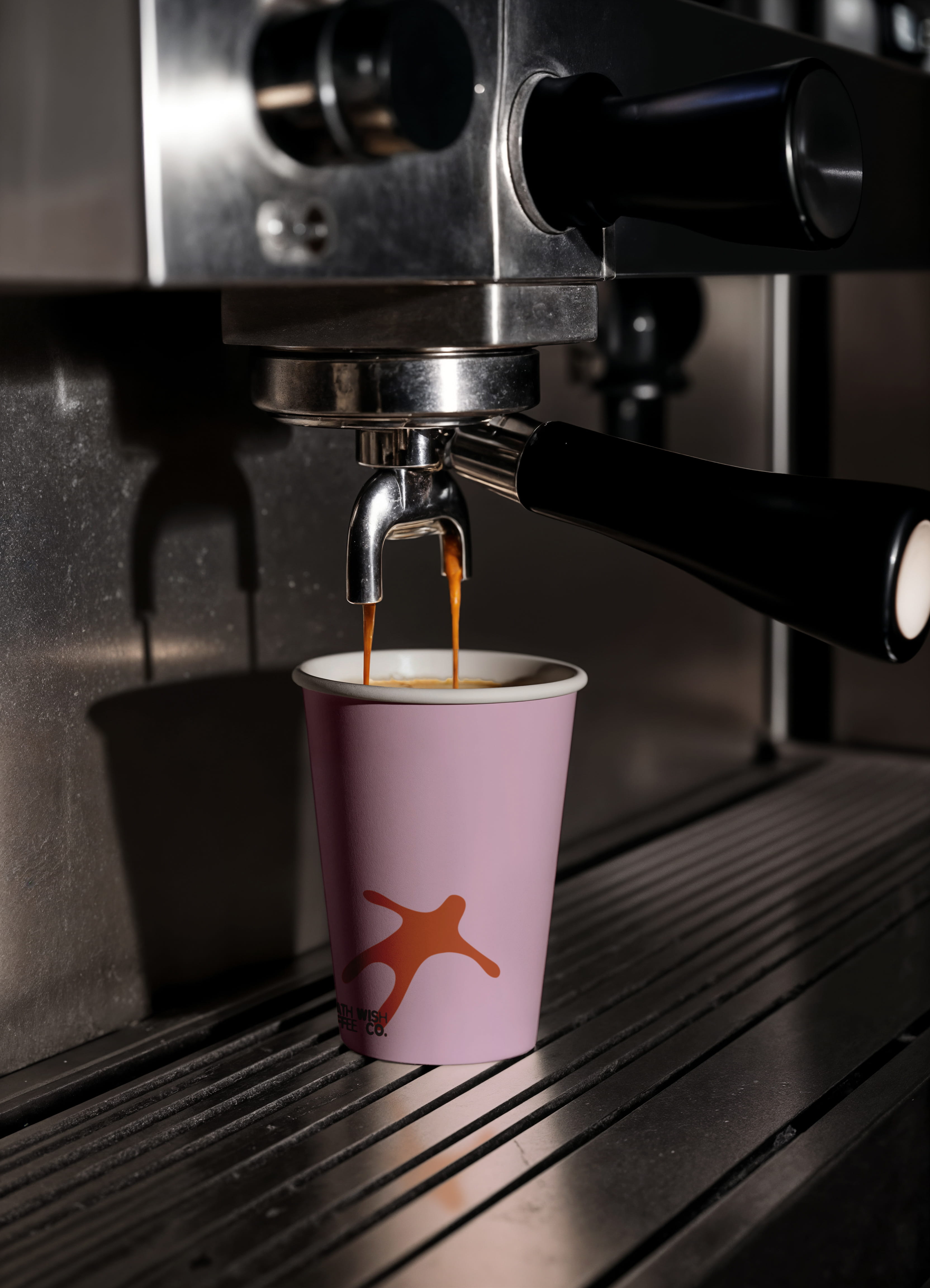 mockup found on magnific -Coffee flows from a metal espresso machine into a pink paper cup with an orange abstract logo.