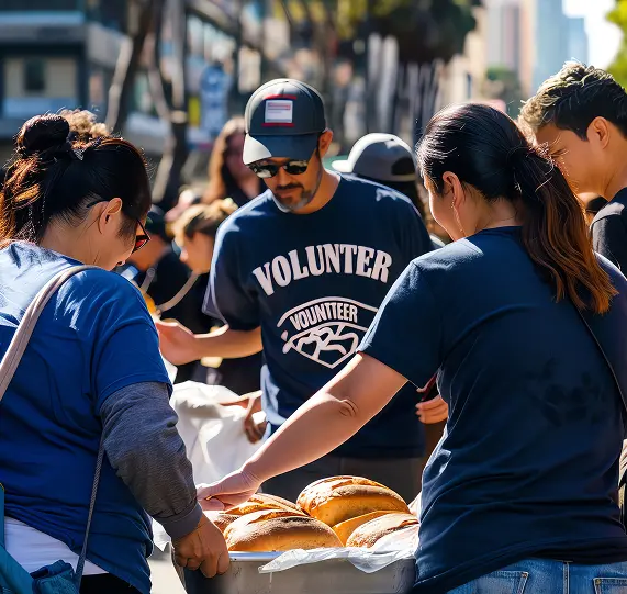 A group of volunteers, wearing matching blue shirts, are gathered around a table with trays of bread rolls. They appear to be serving food to people in a busy urban setting, with buildings and palm trees in the background.