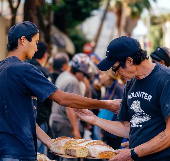 Two volunteers are distributing bread to individuals in a community setting. One volunteer, wearing a black cap and a black shirt, hands a loaf of bread to an older man in glasses and a black shirt that reads 'VOLUNTEER.' Background shows a gathering of people.