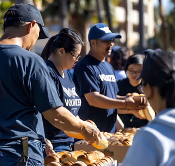 A group of volunteers in matching blue shirts are organizing food items in a community service event. They are standing behind tables with boxes filled with bread rolls, with people participating in the background.