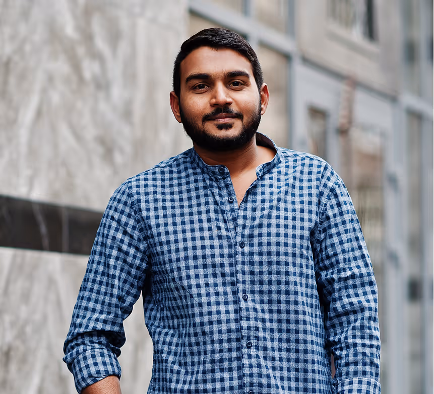 A man with a beard wearing a blue and white checkered shirt stands against a blurred urban background. Bright green digital distortions overlay parts of the image, creating an abstract visual effect.
