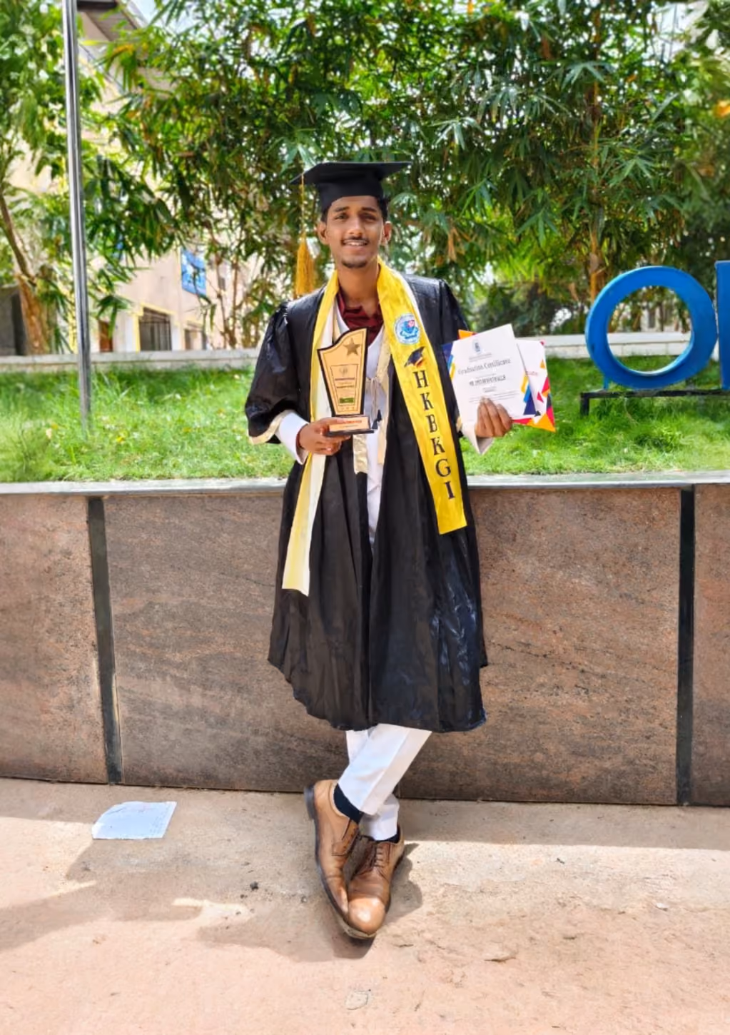 A graduate wearing a black graduation gown and cap holds a trophy in one hand and a diploma in the other. He stands outdoors in front of greenery, with a colorful banner and a stone wall visible in the background.