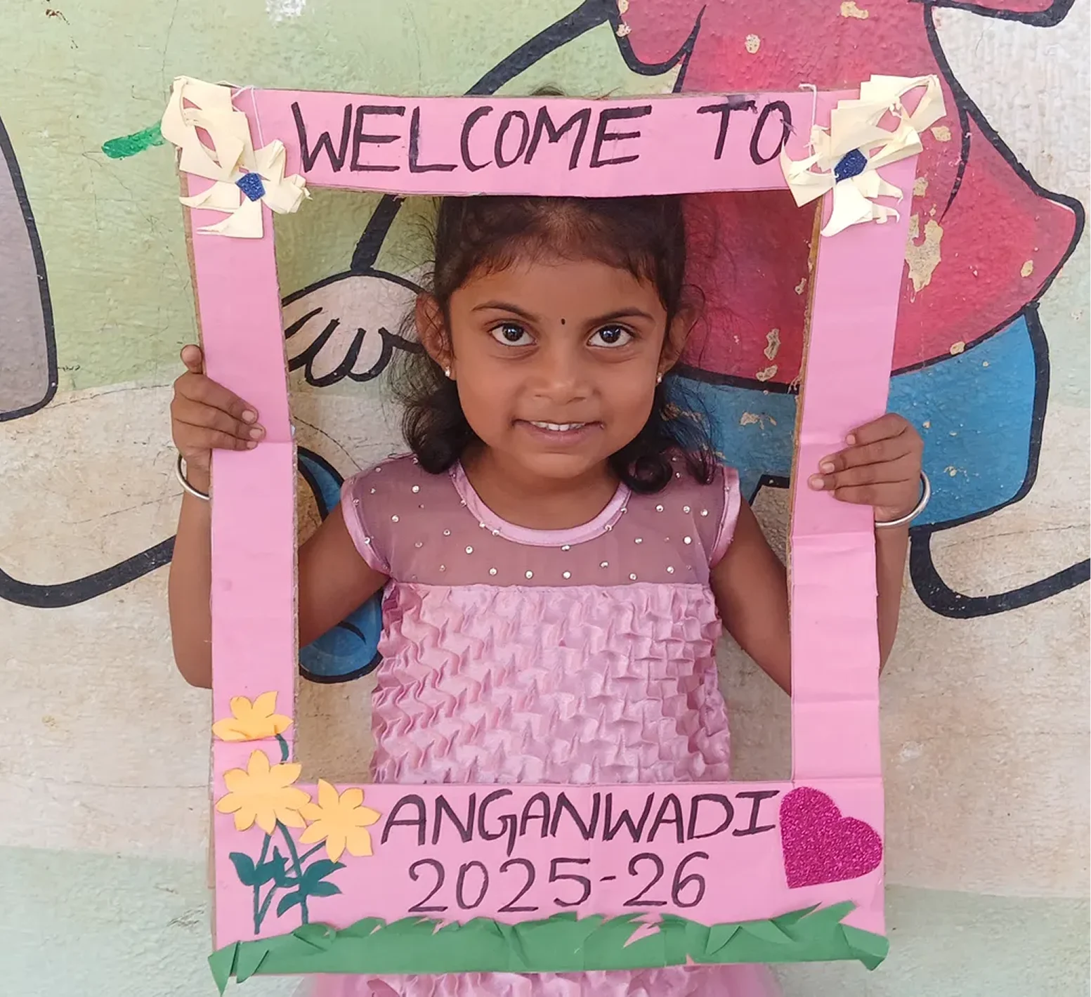 A young girl holding a colorful, hand-made frame with the text 'Welcome to Anganwadi 2025-26' written on it. She is wearing a pink dress and has curly hair. The background features a painted design on the wall.