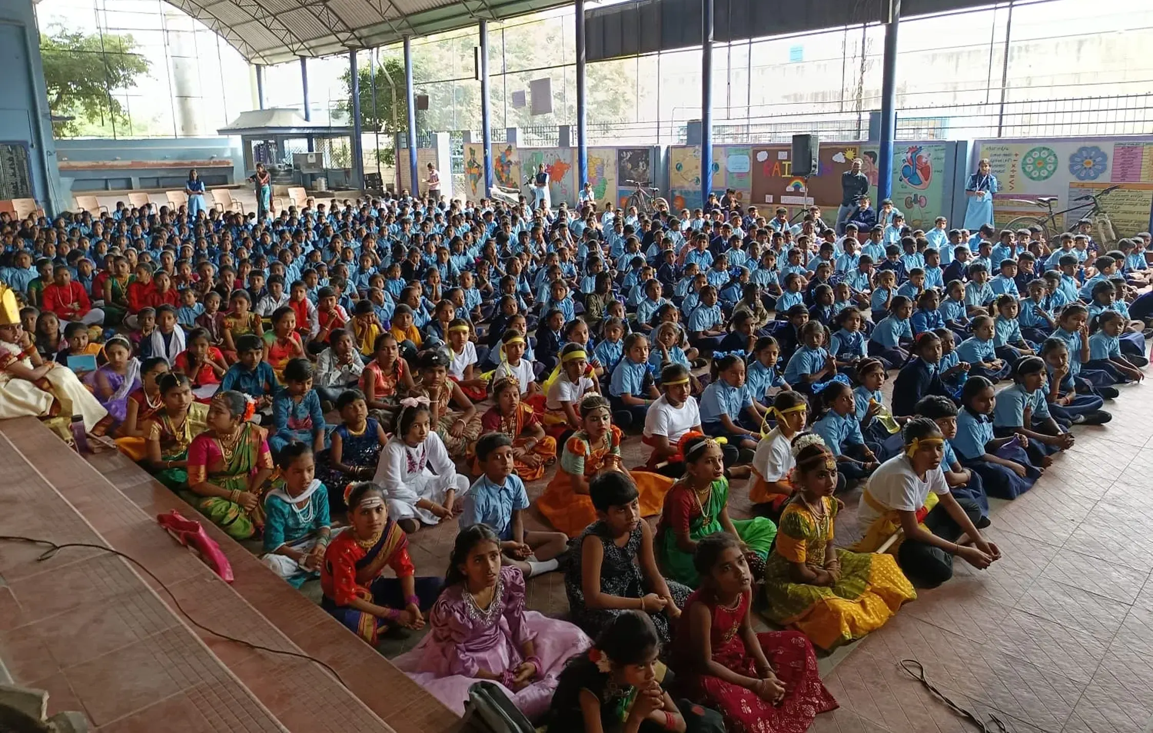 A large gathering of children seated on the floor in an indoor venue. The assembly includes students dressed in colorful traditional attire and school uniforms, with a backdrop of educational decorations and windows allowing natural light.