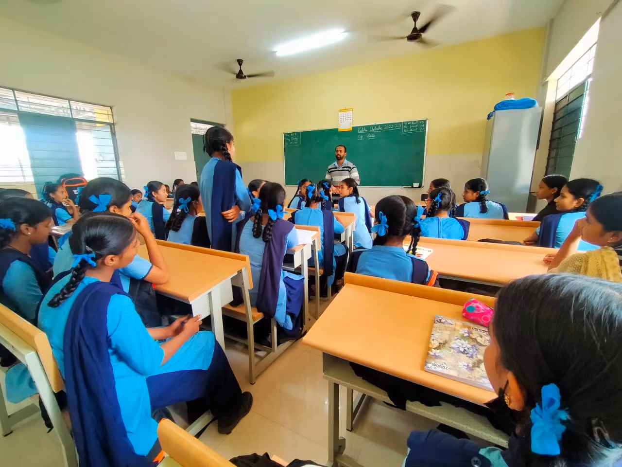A classroom filled with students in blue uniforms sitting at desks. A teacher stands in front of a blackboard while students face him, some raising their hands. The room has large windows, allowing natural light to enter.