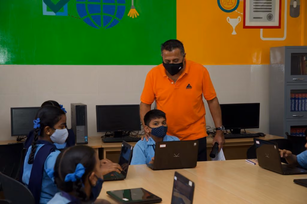 A teacher in an orange polo shirt leans over a young student wearing a blue shirt and a mask, who is sitting at a table with a laptop. In the background, several children in blue uniforms sit with laptops in a classroom with educational wall decorations.
