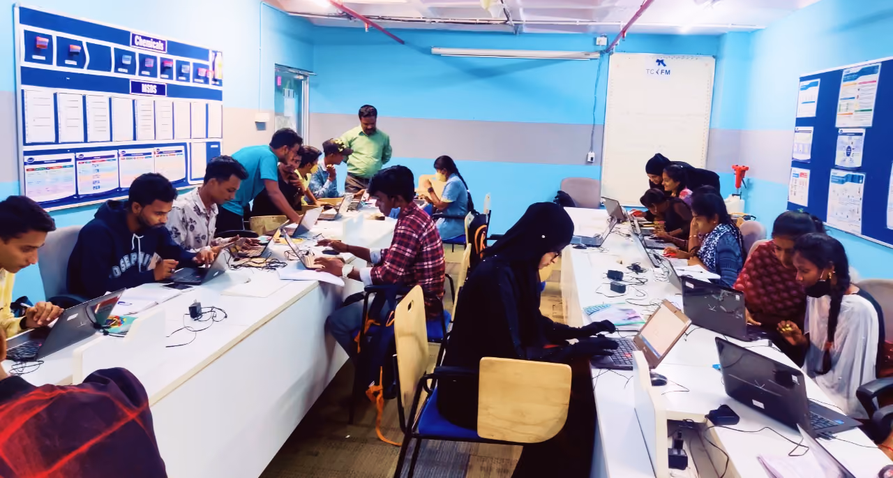 A group of individuals working on laptops at a long table in a brightly lit office space. Some people are seated at the table while others stand nearby, with various electronic devices and papers scattered across the workspace.