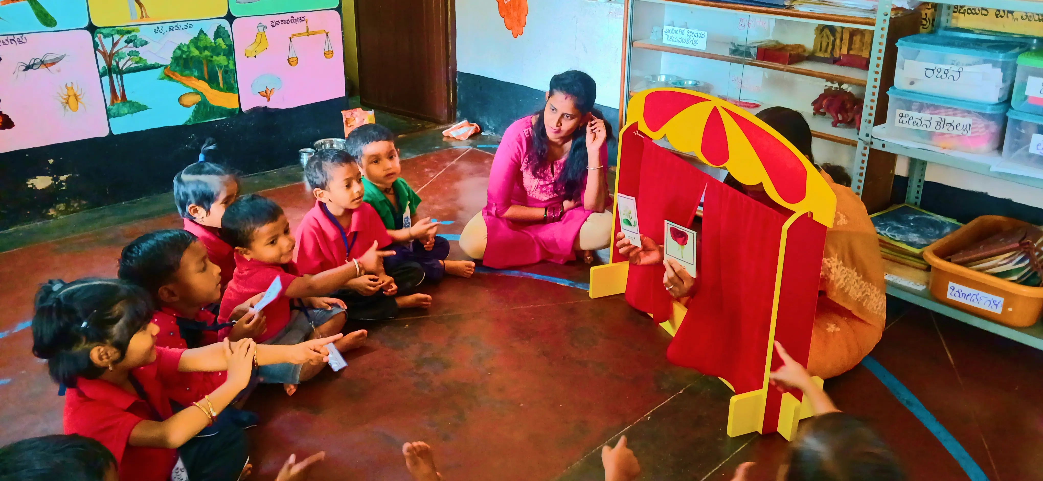 A group of young children in red shirts sit on the floor, engaged in a storytelling session. A woman in a pink outfit operates a puppet theater with colorful curtains, while the children eagerly point and watch attentively.