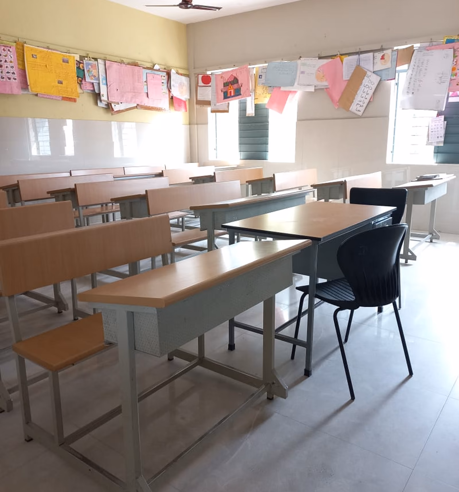Empty classroom with wooden desks and chairs, student artwork hanging on strings near windows.