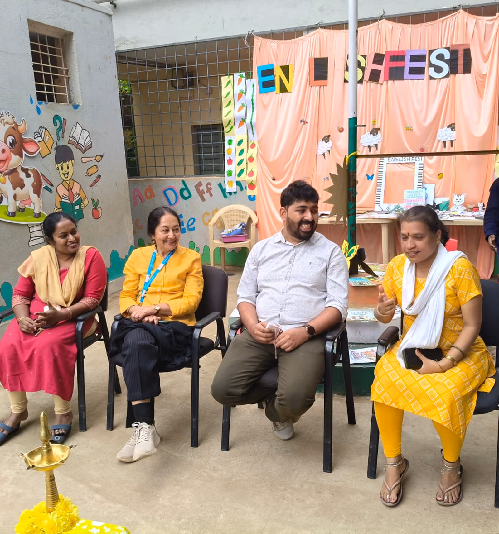 Four adults seated outdoors in front of colorful educational wall and English Fest display, smiling and talking.