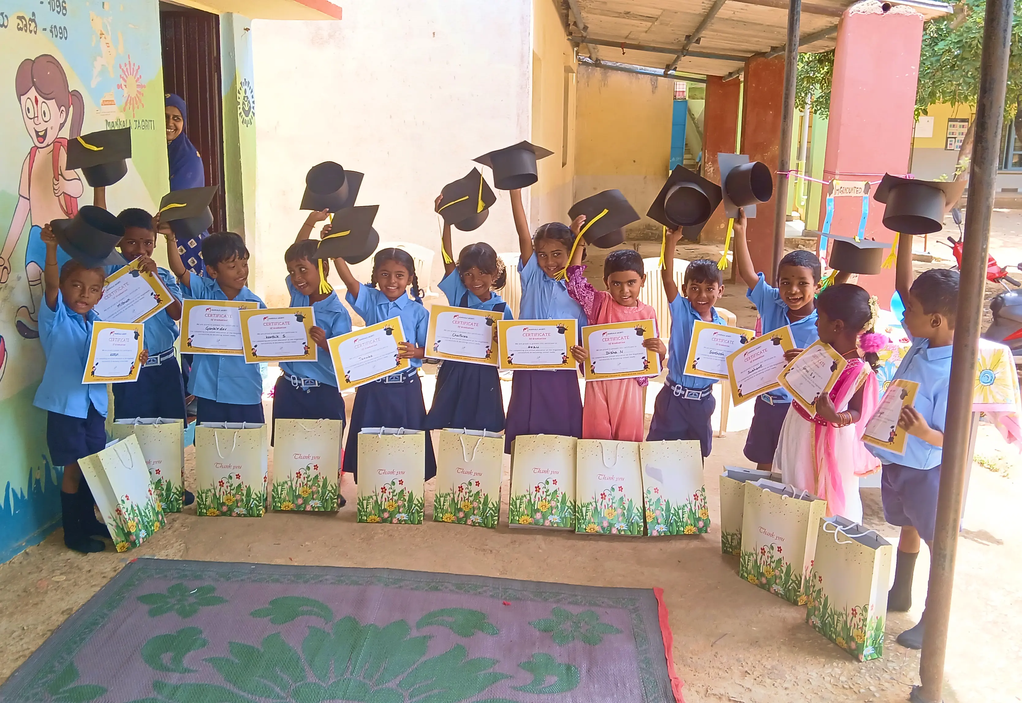 Children in blue uniforms holding certificates and raising graduation caps, smiling at an outdoor event.