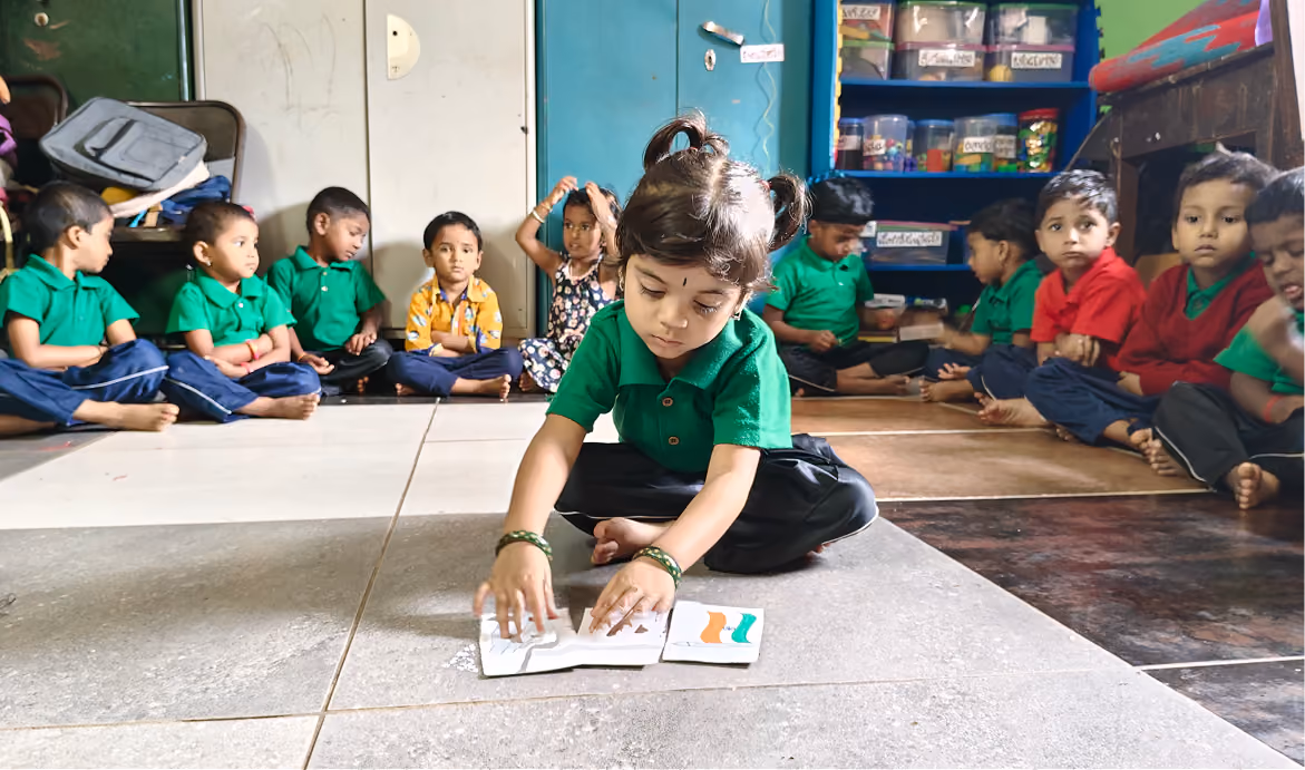 Group of young children sitting in a circle on floor drawing and showing artwork in colorful classroom.