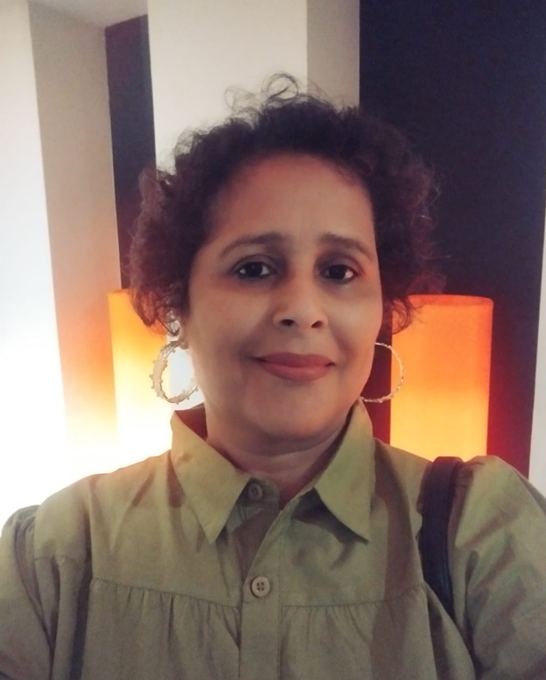 Woman smiling, wearing a gold embroidered saree with matching earrings and a decorative bindi.