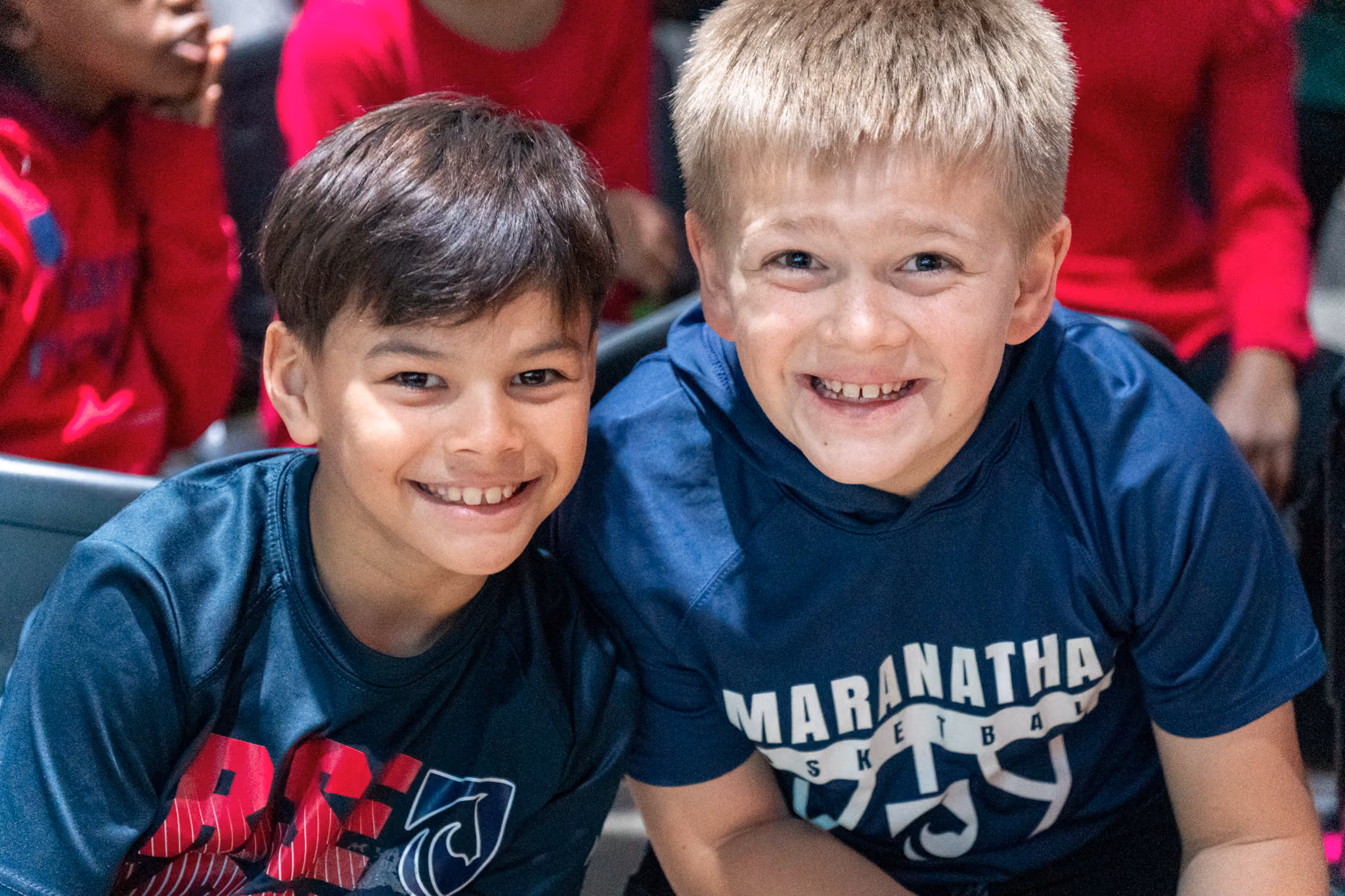 High school student in a grey Mustangs sweatshirt smiling while giving a piggyback ride to a younger elementary student.