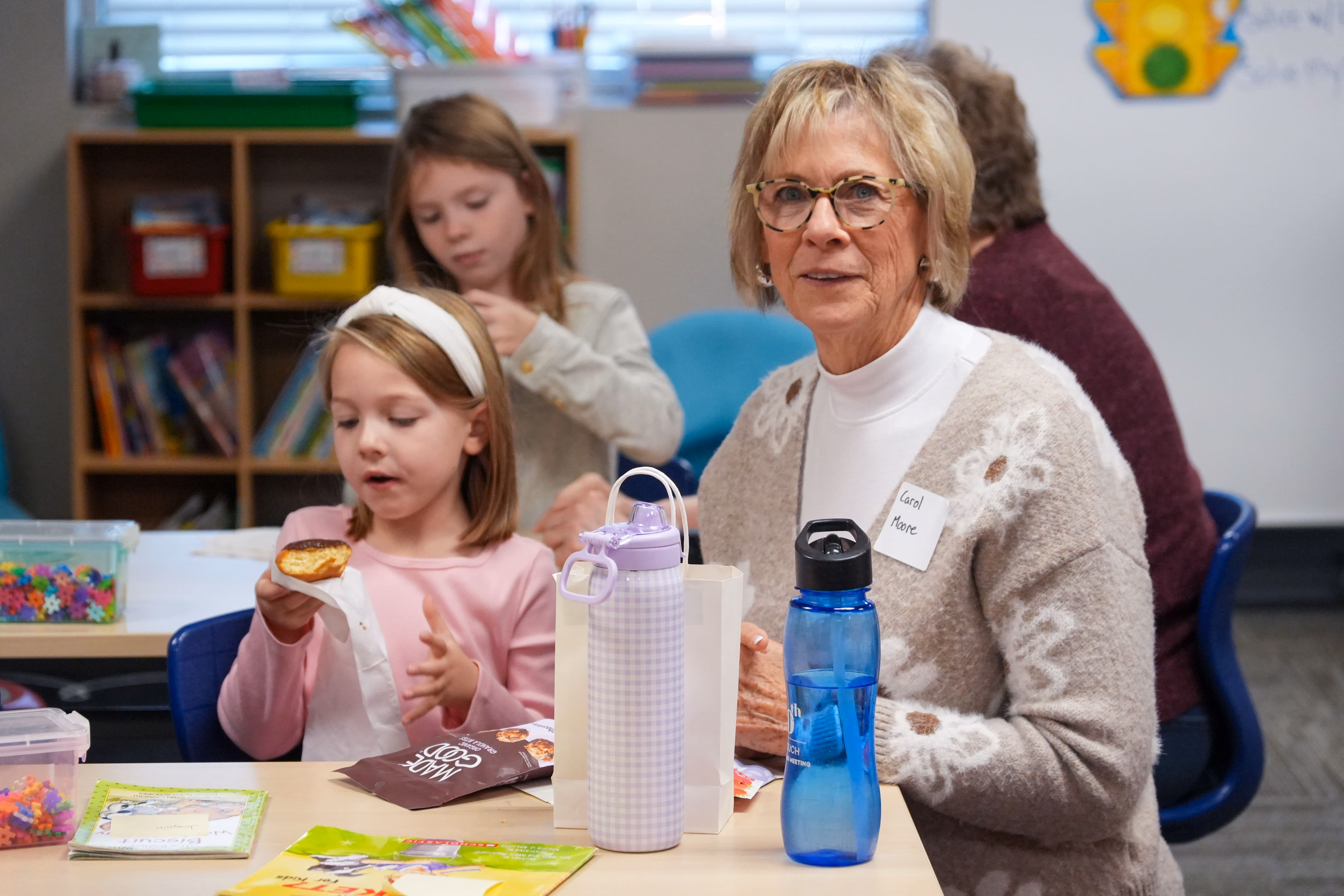 A grandmother sitting at a classroom table alongside young students enjoying a snack.