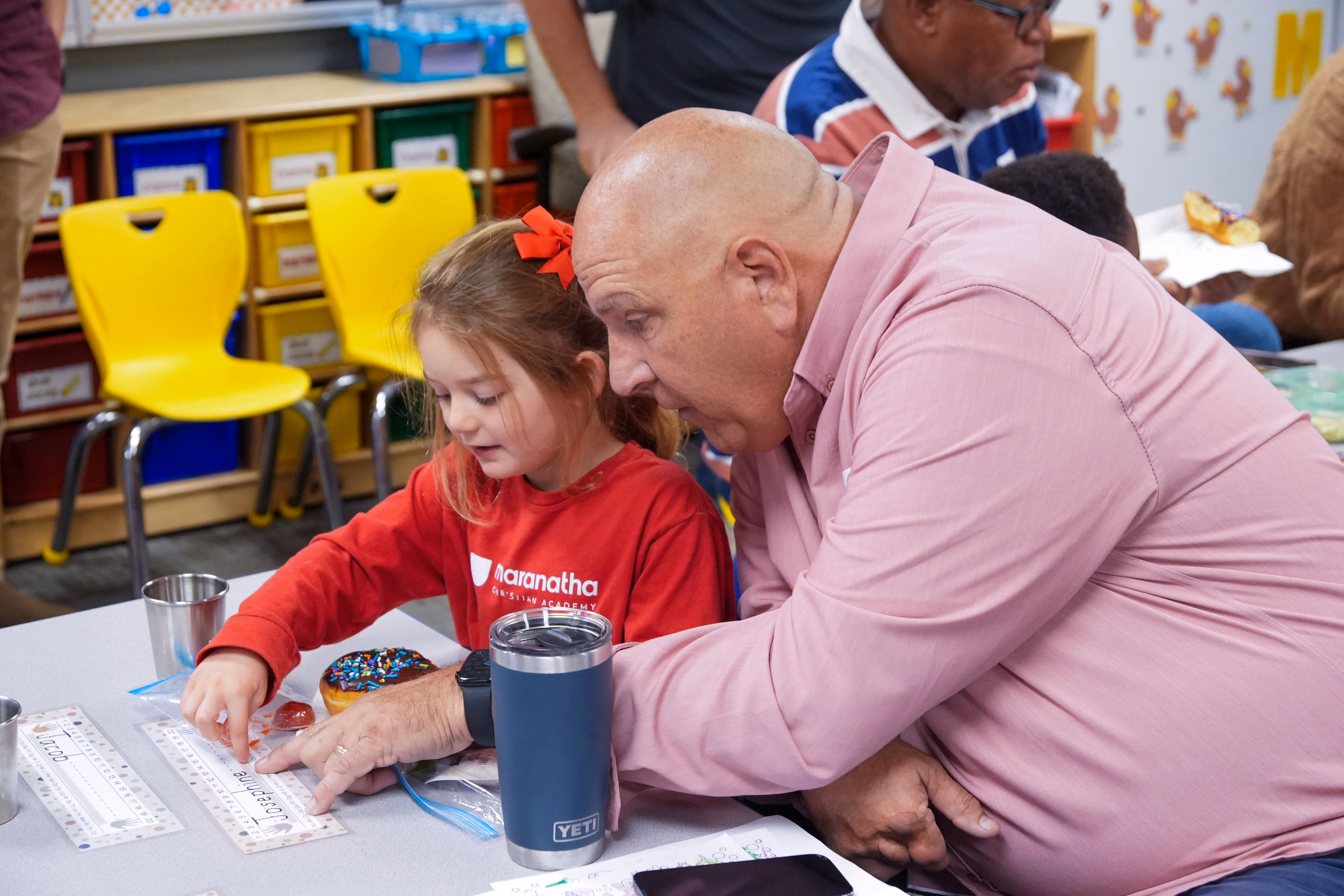 A grandfather and his young granddaughter, who is wearing a red Maranatha Christian Academy shirt, looking closely at something together.