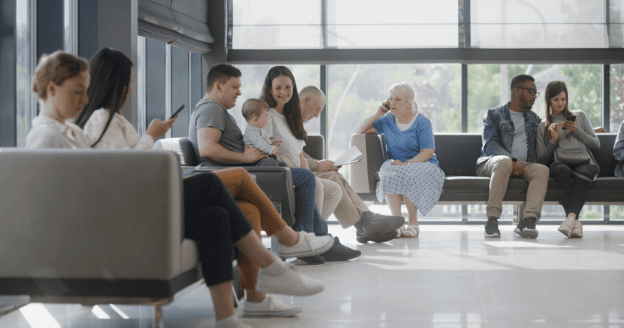 Diverse group of adults and children waiting in bright, contemporary healthcare facility with large windows and modern seating.