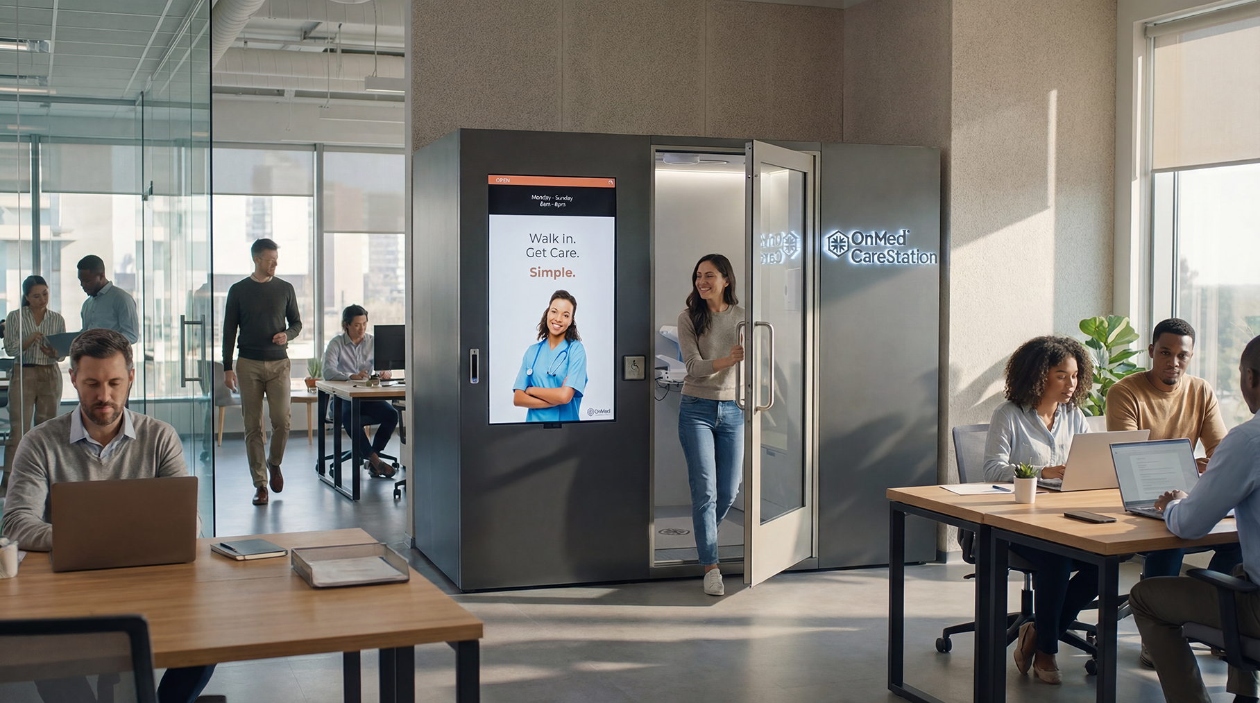 OnMed CareStation self-service health kiosk deployed in open office with employees working at desks, providing on-demand employer healthcare access.