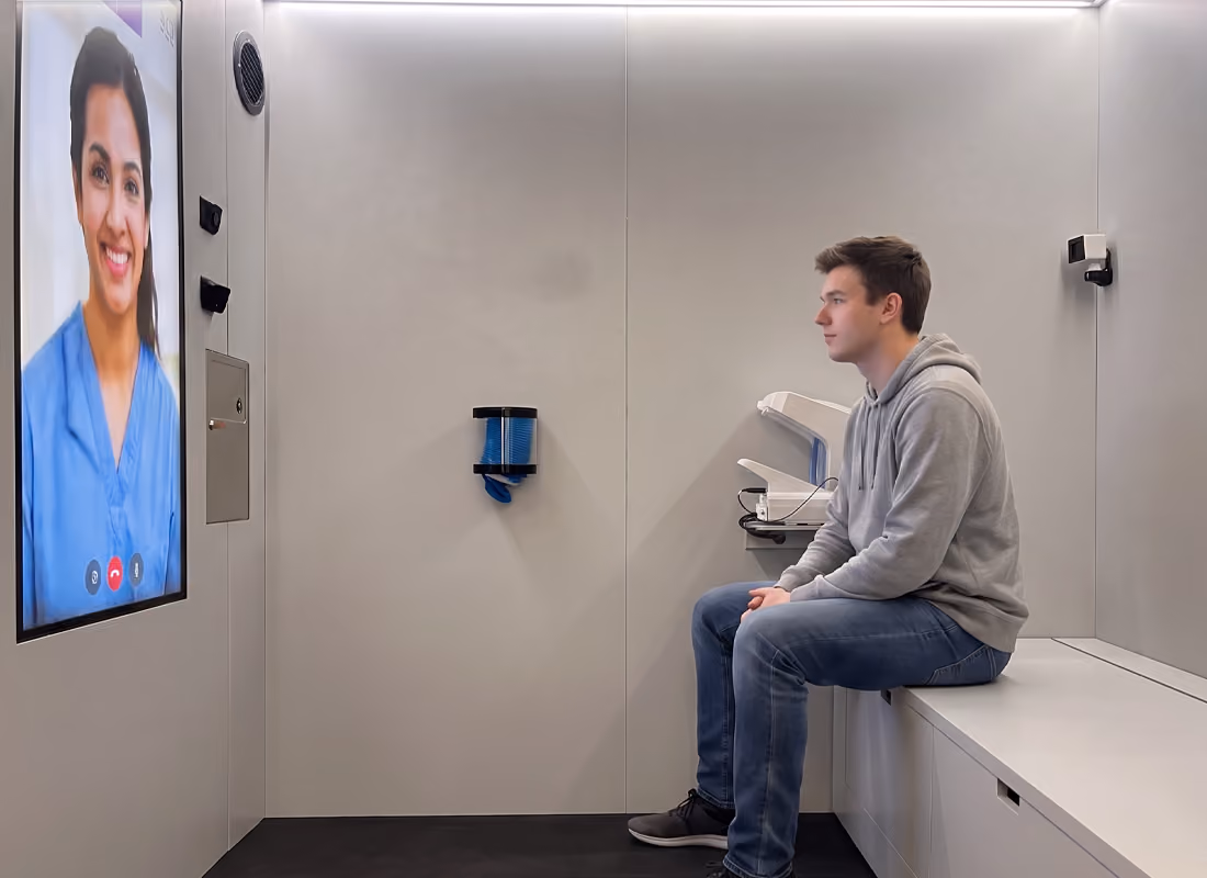 Young man in a hoodie seated inside an OnMed CareStation™, speaking with a clinician on the video screen