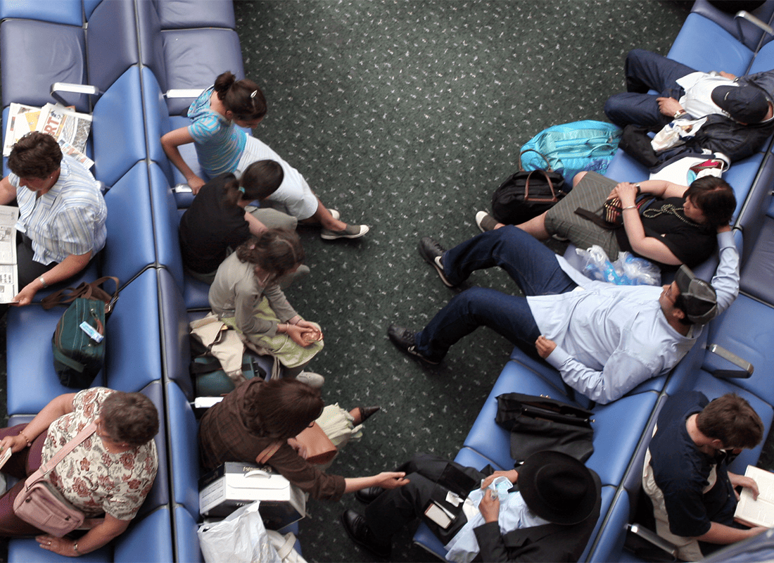 Overhead view of travelers with luggage sitting and lying across rows of blue lounge seating in a crowded waiting area.