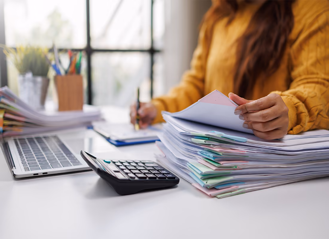 Person in a yellow sweater sorting through a large stack of paperwork with a calculator and laptop nearby