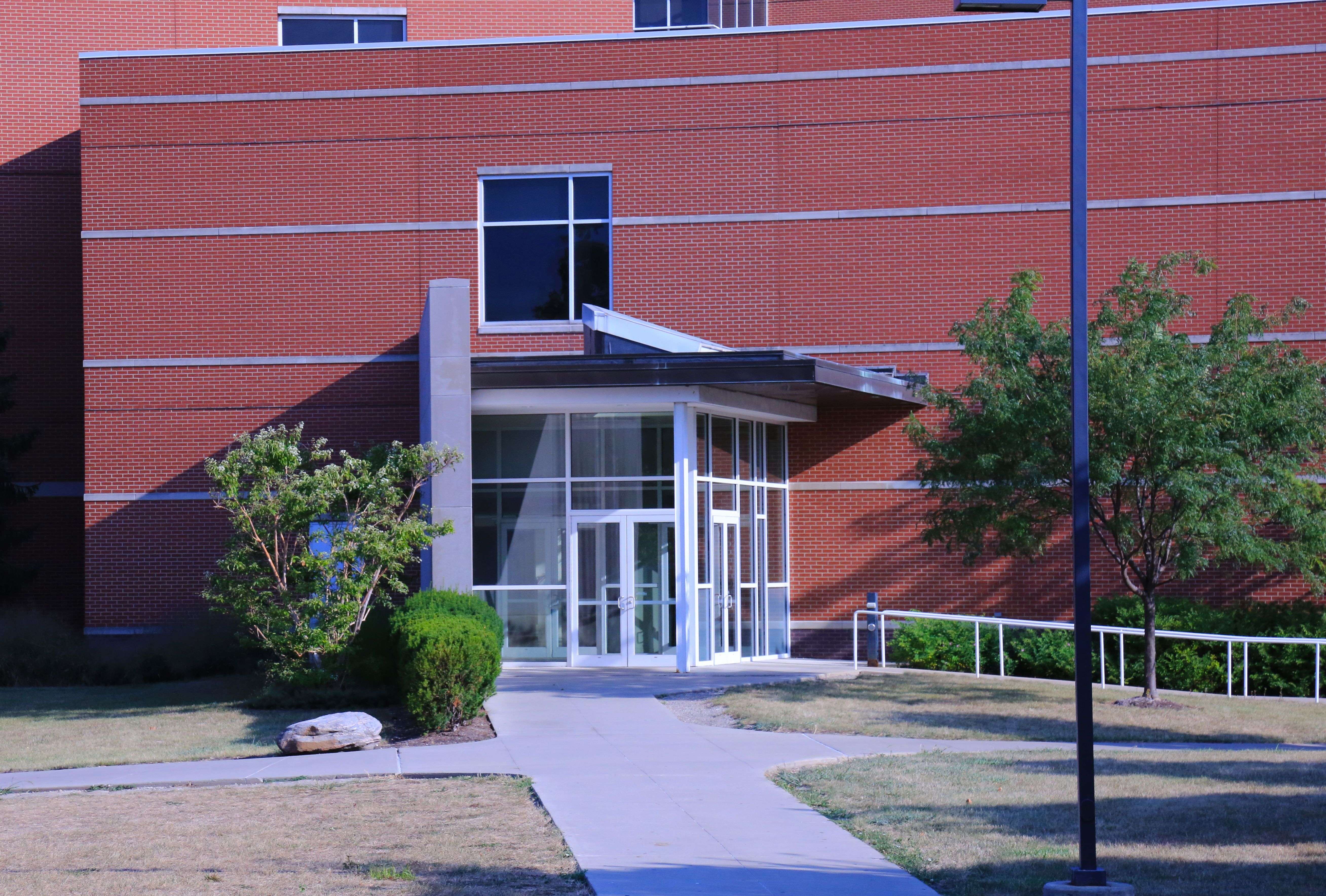 Red brick institutional building entrance with glass double doors, covered awning, and landscaped walkway.