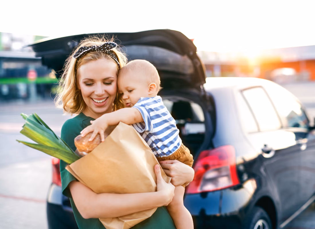 Woman smiling while holding a bag of groceries and her toddler next to a car