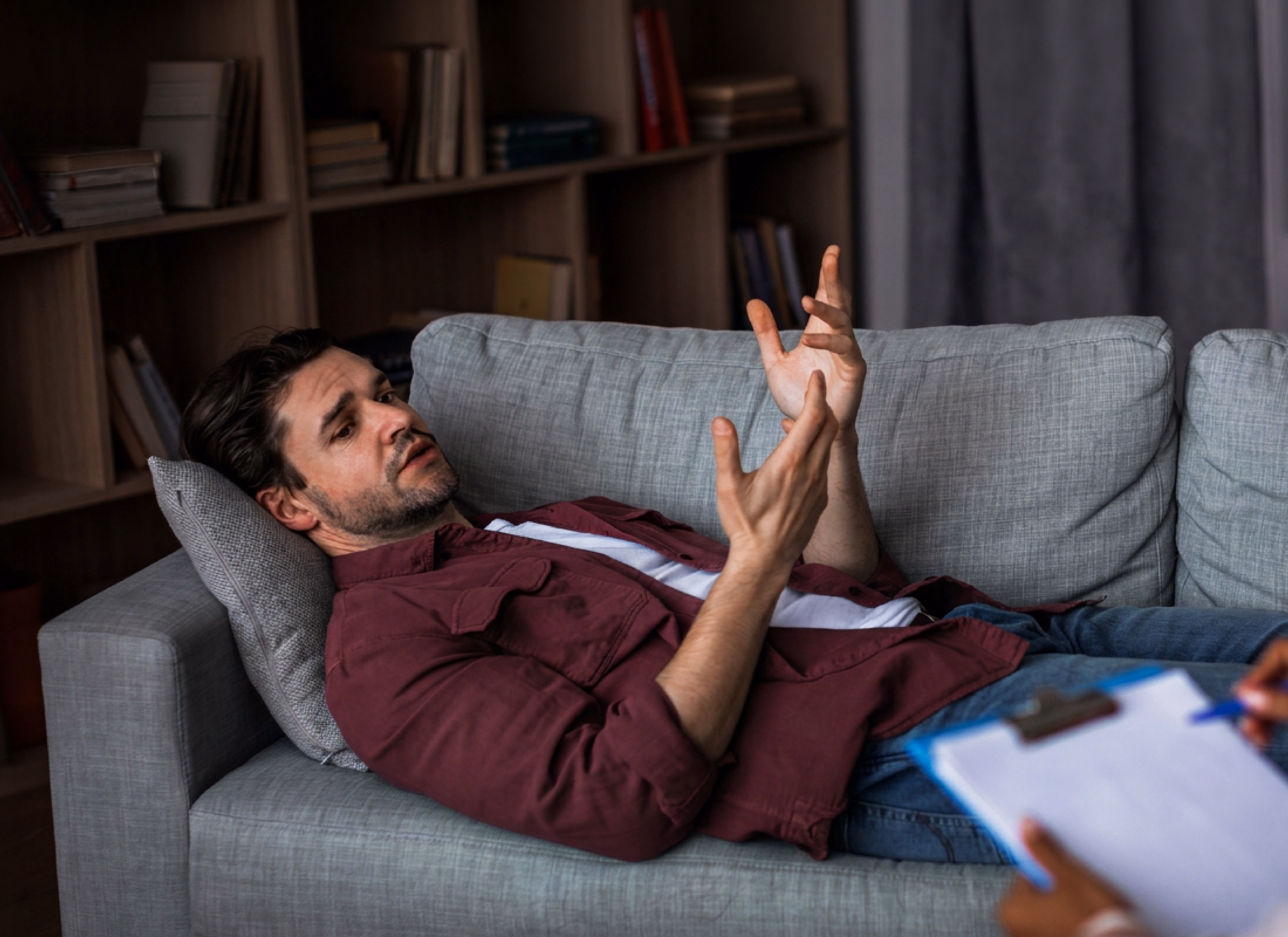 Man reclining on a gray sofa, gesturing with his hands as he speaks, with a clipboard visible in the foreground