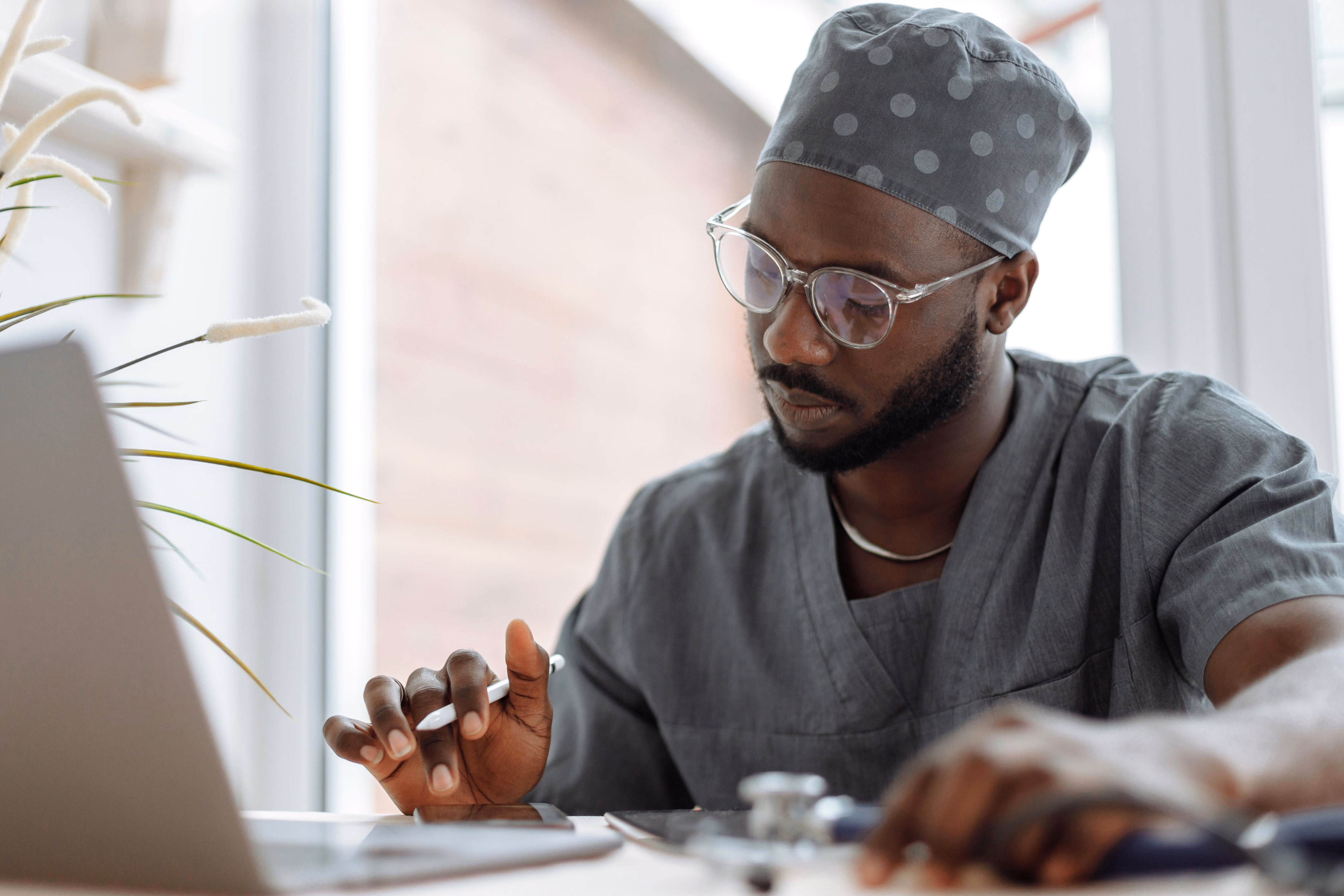 Clinician in gray scrubs and a surgical cap reviewing information on a tablet with a stylus at a desk with a laptop and stethoscope
