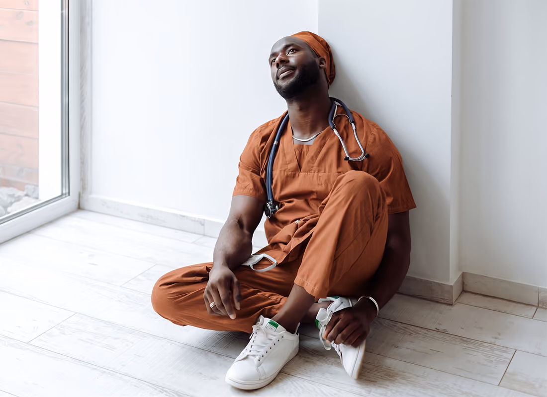 Healthcare worker in rust-colored scrubs with a stethoscope sitting on the floor against a wall, looking upward in a thoughtful pause