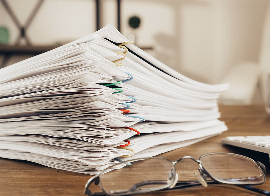 Tall stack of paperwork held with colored paper clips on a wooden desk next to a pair of reading glasses