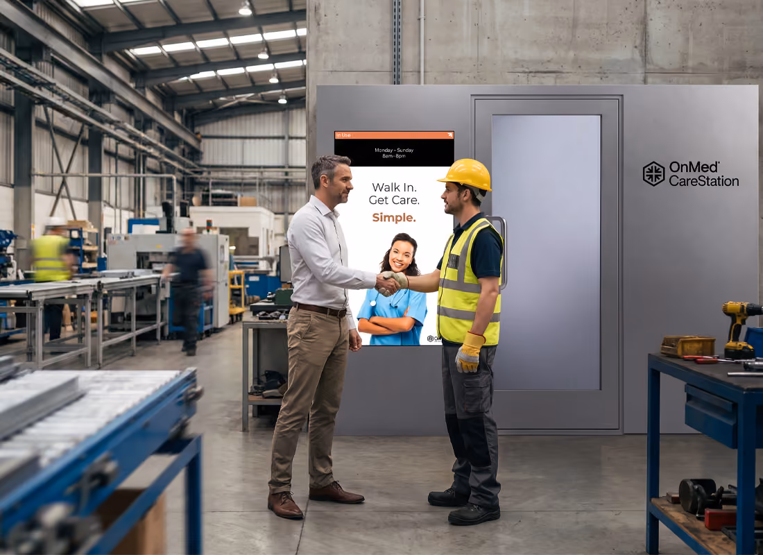 Two men shaking hands in front of an OnMed CareStation® with a "Walk In. Get Care. Simple." screen inside a manufacturing facility
