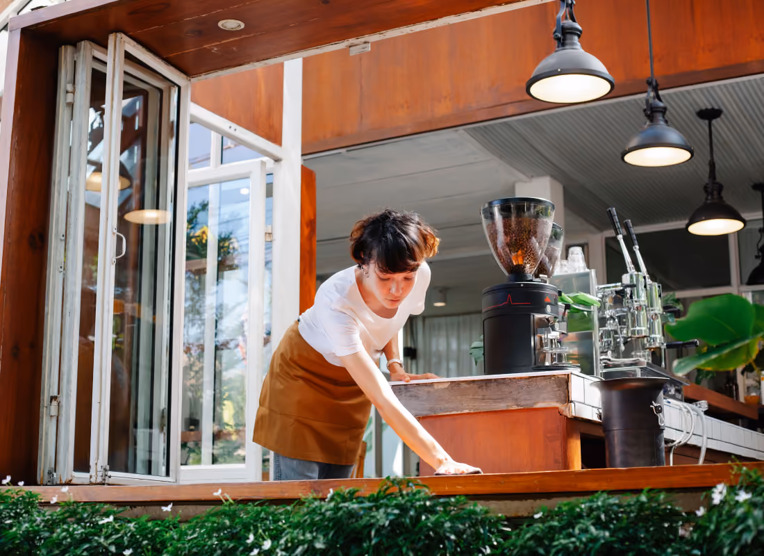 Cafe worker in an apron wiping down a counter beside a coffee grinder and espresso machine at an open-air coffee shop