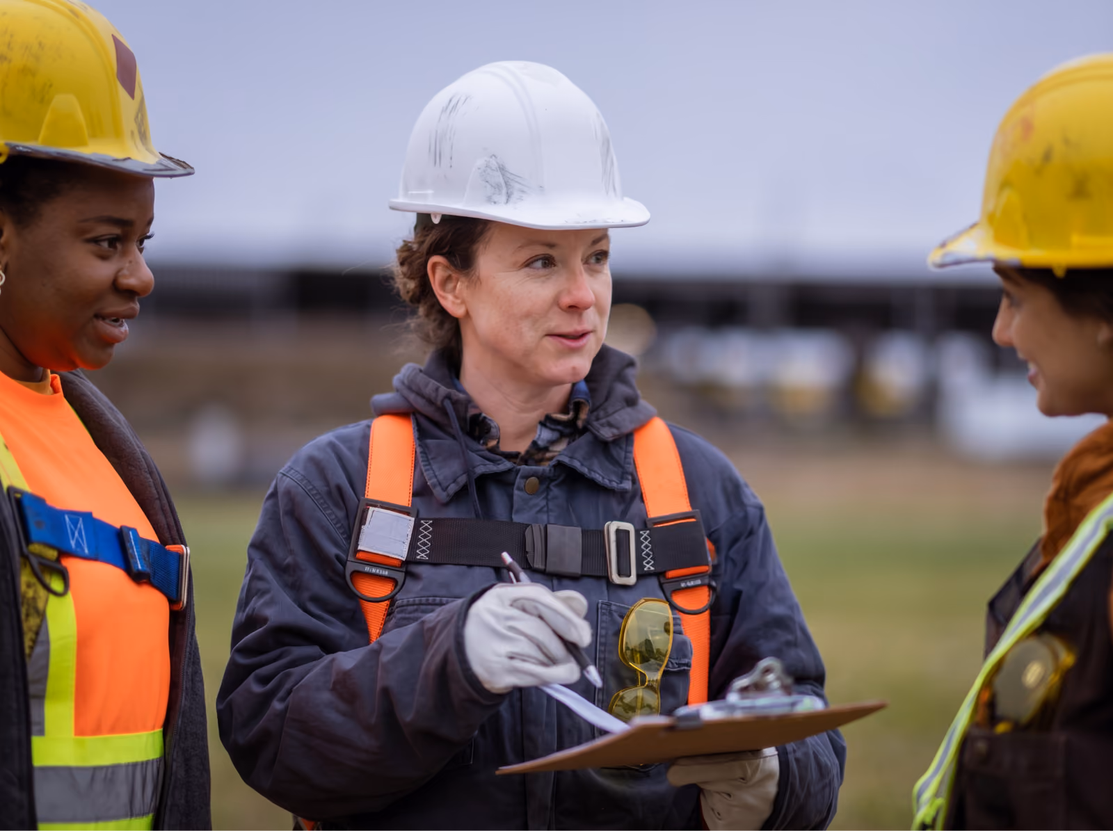 Three workers in hard hats and safety vests talking on a job site as one reviews a clipboard
