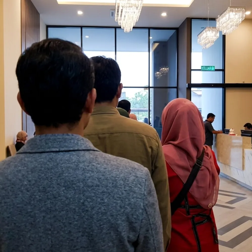 People standing in line at a modern reception counter with chandeliers and large windows.