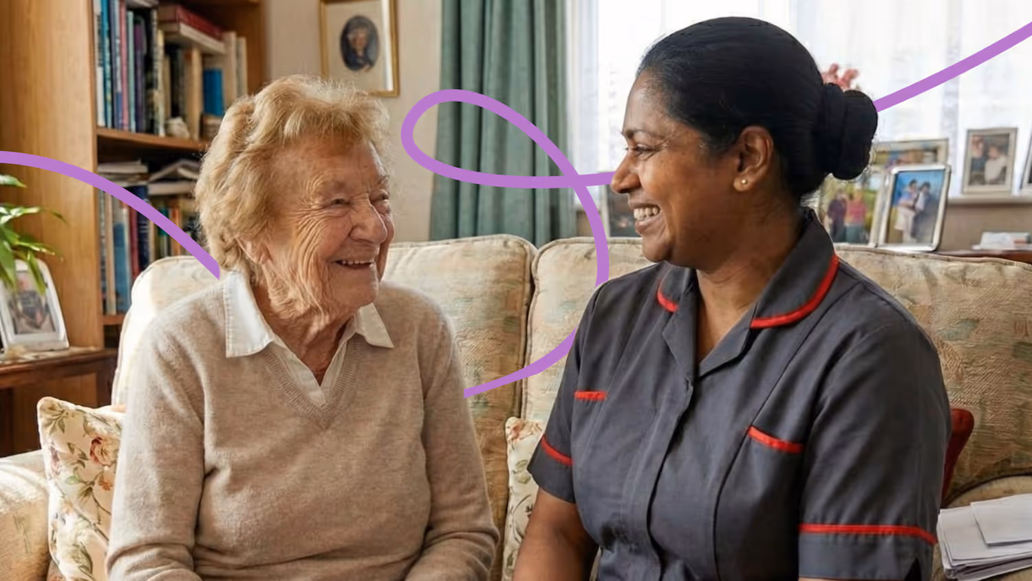 A Routes Healthcare carer sits with an elderly female client on a sofa in her home, both sharing a genuine laugh together.