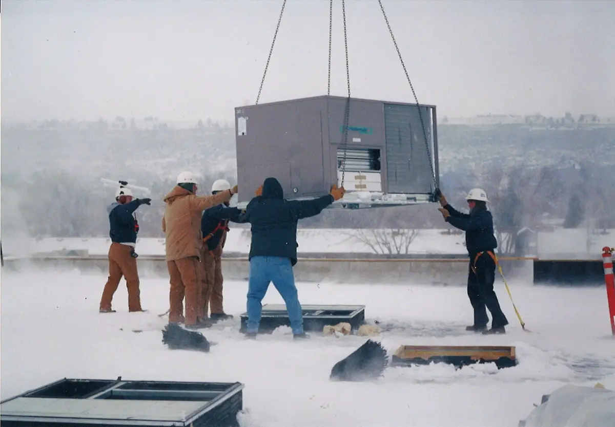Five workers install a rooftop HVAC unit on a snowy day while wearing winter gear.