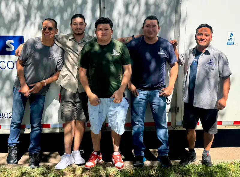 Five men standing side by side outdoors, smiling in front of a white truck with company logos.