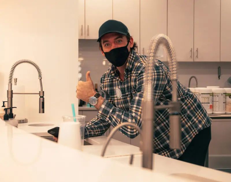 A plumber wearing a plaid shirt and black face mask giving thumbs up in a modern kitchen near a sink.