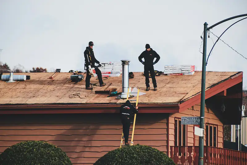 Three workers on a residential roof installing shingles with ladders and roofing materials.