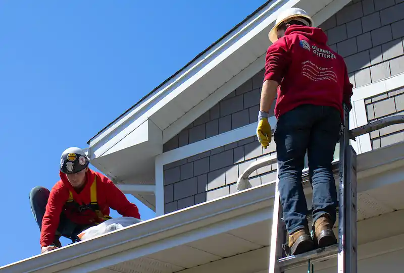 Two workers wearing helmets and red jackets installing or repairing gutters on a house roof.