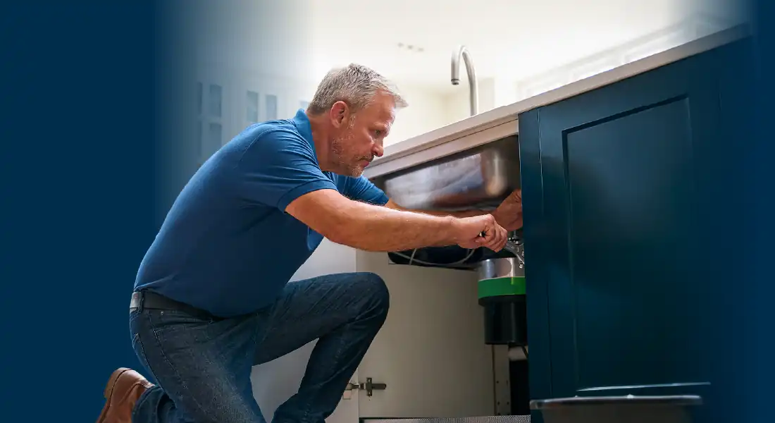 Man in blue shirt kneeling and fixing plumbing under kitchen sink with blue cabinets.